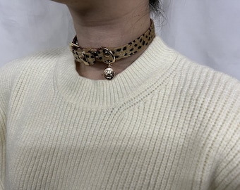 May include: A brown and black leopard print choker necklace with a gold charm. The necklace is worn by a person wearing a white sweater.
