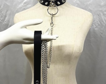 Può includere: Choker in pelle nera con borchie argentate e un grande anello di metallo. Sono attaccati un cinturino nero abbinato e una catena argentata. Il choker è esposto su un manichino.