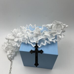 May include: White lace choker with a black cross pendant. The choker is made of delicate lace and has a ruffled edge. The cross pendant is large and ornate.