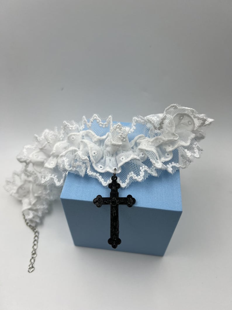 May include: White lace choker with a black cross pendant. The choker is made of delicate lace and has a ruffled edge. The cross pendant is large and ornate.