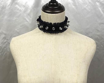 Könnte beinhalten: Ein schwarzes Choker-Halsband mit silberfarbenen Stacheln und einem Rüschenbesatz. Die Halskette wird auf einer Schaufensterpuppe präsentiert. Ein Accessoire im Gothic- oder Punk-Stil.