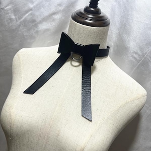 Könnte beinhalten: Eine schwarze Leder-Halskette mit Schleife und langen, herabhängenden Riemen. Die Halskette hat einen silberfarbenen Metallring in der Mitte der Schleife. Die Halskette wird auf einer Schaufensterpuppe präsentiert.
