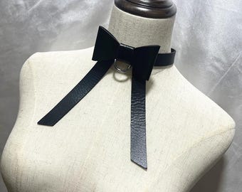 May include: A black leather choker necklace with a bow and long, dangling straps. The choker features a silver-colored metal ring at the center of the bow. The necklace is displayed on a mannequin.