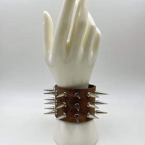 May include: A wide brown leather cuff bracelet featuring three horizontal rows of sharp, silver-toned metal spikes. The leather has a distressed, textured finish, and the spikes are arranged in a uniform pattern around the entire band.