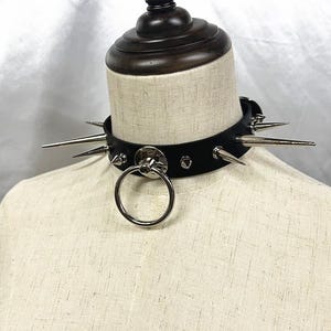 Może przedstawiać: Czarny skórzany choker ze srebrnymi kolcami i dużym metalowym pierścieniem. Choker jest nabijany wieloma srebrnymi kolcami i małymi ćwiekami. Pierścień jest przymocowany do przodu chokera.