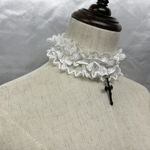 May include: White lace choker with a black cross pendant. The choker is made of delicate lace and has a ruffled design. The cross pendant is simple and elegant.