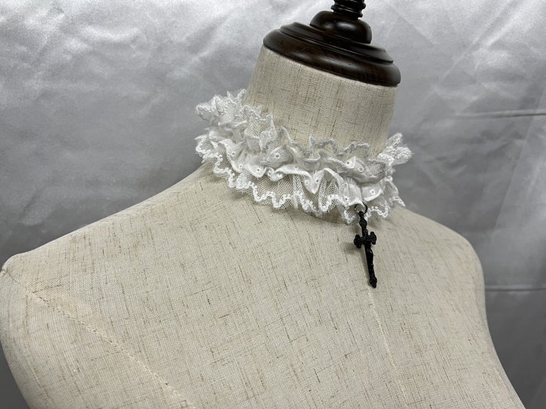 May include: White lace choker with a black cross pendant. The choker is made of delicate lace and has a ruffled design. The cross pendant is simple and elegant.