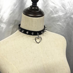 May include: Black choker necklace with silver-colored spikes and a heart pendant. The necklace is displayed on a mannequin. The choker is made of a black material and has a row of silver spikes around the neck.