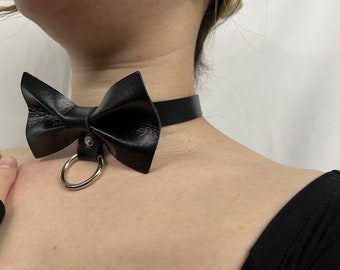 May include: A black leather choker with a bow tie and a silver ring. The choker is worn by a person with a black shirt.