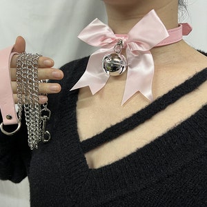 May include: A pink satin bow choker with a silver bell charm. The choker is worn on a person wearing a black sweater.
