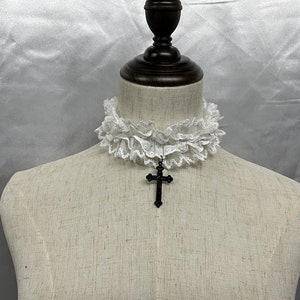 May include: White lace choker with a black cross pendant. The choker is made of delicate lace and has a ruffled edge. The cross pendant is simple and elegant.