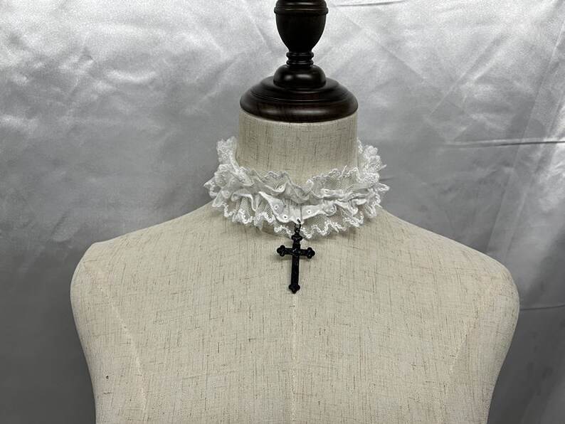 May include: White lace choker with a black cross pendant. The choker is made of delicate lace and has a ruffled edge. The cross pendant is simple and elegant.