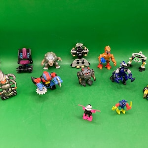 May include: A collection of colorful toy robots and figures, including a silver robot with horns, a red and blue robot with a saw blade, and a yellow and black vehicle. The toys are arranged on a green surface.