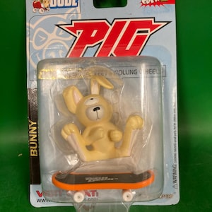 May include: A Tech Deck Dude toy featuring a yellow bunny figure on a miniature skateboard. The skateboard has an orange stripe and white wheels. The packaging includes the text "PIG" and "SKATE CREW 2". Ages 9 & up.