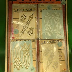 May include: Vintage decal sheets with various designs, including abstract shapes and lines. The packaging features the word "DECALS" in large letters, along with the phrase "It's easy to decorate with." The decals are designed for bikes, windshields, boats, and lamps.
