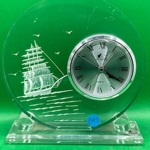 May include: A clear glass clock with a white sailboat design and a silver clock face with black Roman numerals. The clock face reads "CAT Designs Quartz".
