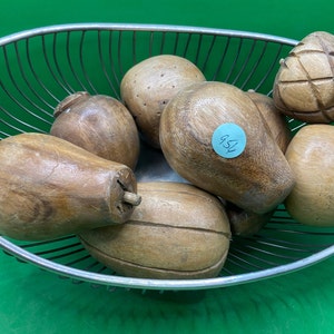 May include: A metal wire basket filled with various wooden decorative fruits. The fruits are in shades of brown and tan, with different shapes and textures. One fruit has a carved geometric pattern. A blue sticker with the number "952" is visible.