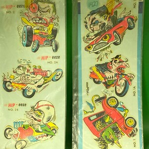 May include: Two vintage decal sheets featuring cartoon hot rod characters and vehicles. The decals are in clear plastic packaging. The text "HIP-eeze" and numbers are visible on the decals. The background is a light blue color.