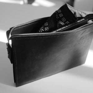 Puede incluir: Una bolsa de cuero negro con cremallera con un pañuelo negro y blanco en su interior. La bolsa tiene una cremallera y una solapa con cierre de presión.