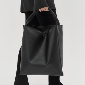 Puede incluir: Un bolso tote grande de cuero negro con un diseño holgado. El bolso tiene una abertura ancha y dos asas largas.