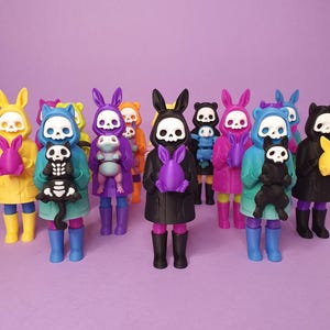 May include: A group of figurines with skull faces and bunny ears, dressed in coats and holding toys. The figurines are in a range of colours, such as yellow, blue, purple, and black, and are positioned on a purple surface.