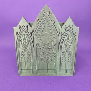 May include: A sage green container in a gothic style, featuring an intricate facade. The design includes arched windows, decorative patterns, and a pointed crown. The container is made of a solid material and rests on a purple surface.