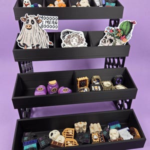 May include: A black tiered display shelf with four levels of compartments containing various small decorative items, including miniature skulls, potion bottles, candles, and stickers featuring a ghost cow with the text MOOOOO I MEAN BOOOOO.
