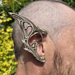 May include: A metallic gold-toned ear cuff featuring an intricate, pointed, and elongated design that extends upwards and downwards along the outer edge of the ear. The accessory has a textured, layered appearance with cut-out details and a curved structure.