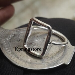 Peut inclure: Une bague en argent avec un design rectangulaire ouvert. La bague est pos&eacute;e sur une surface argent&eacute;e orn&eacute;e. Le texte "Kpringstore" est visible. Un bijou minimaliste.