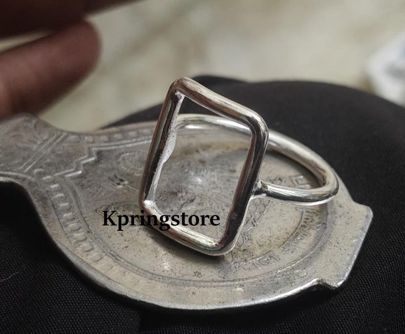 Peut inclure: Une bague en argent avec un design rectangulaire ouvert. La bague est pos&eacute;e sur une surface argent&eacute;e orn&eacute;e. Le texte "Kpringstore" est visible. Un bijou minimaliste.
