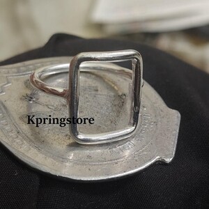 Peut inclure: Une bague argent&eacute;e avec un design rectangulaire. La bague a une esth&eacute;tique simple et moderne. La bague est pr&eacute;sent&eacute;e sur une surface argent&eacute;e. Le texte "Kpringstore" est visible sur la surface.
