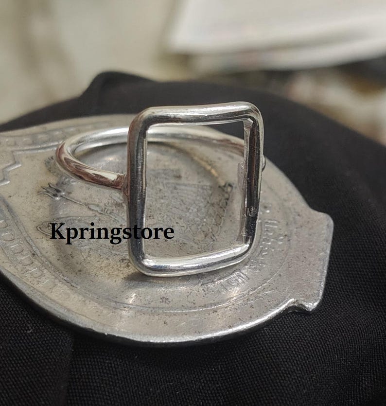 Peut inclure: Une bague argent&eacute;e avec un design rectangulaire. La bague a une esth&eacute;tique simple et moderne. La bague est pr&eacute;sent&eacute;e sur une surface argent&eacute;e. Le texte "Kpringstore" est visible sur la surface.