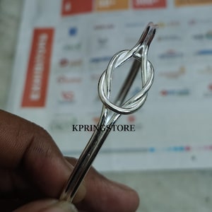 Könnte beinhalten: Ein silberfarbenes Armreif-Armband mit einem Knoten-Design. Das Armband besteht aus glänzendem Metall und hat einen schlichten, eleganten Stil. Der Text "KPRINGSTORE" ist unten im Bild zu sehen.