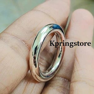 Może przedstawiać: Błyszczący srebrny pierścionek o gładkiej, zaokrąglonej powierzchni. Pierścionek jest pokazany na palcu.