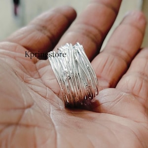 May include: A silver ring with a unique, intricate design that resembles a woven or tangled wire. The ring is wide and appears to be made of multiple strands of silver wire.