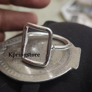 Peut inclure: Une bague en argent avec un design rectangulaire ouvert. La bague est pos&eacute;e sur une surface argent&eacute;e avec le texte "Kpringstore". La bague a un anneau fin et la forme rectangulaire est le point central.