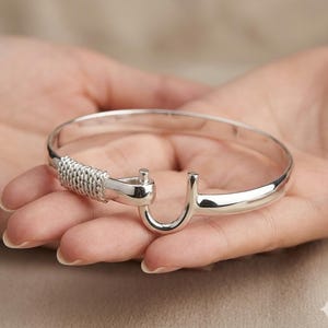 May include: A polished silver-tone bangle bracelet featuring a hook-and-eye style clasp mechanism. One side of the hook is wrapped in a textured, rope-like silver metal detail, creating a nautical-inspired aesthetic with a smooth, reflective finish.