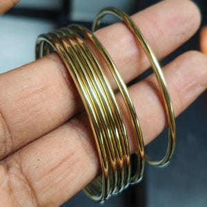 Op de afbeelding: Een set van zes dunne, gepolijste armbanden van goudkleurig metaal. De armbanden hebben een glad, afgerond oppervlak met een reflecterende afwerking. Deze cirkelvormige armbanden zijn gestapeld en hebben een consistente gele metaalkleur.