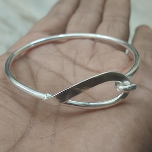 May include: A silver bangle bracelet with an infinity symbol design. The bracelet has a smooth, polished finish and a simple, elegant style. The infinity symbol is formed by a flat, curved piece of metal that connects to the circular band.