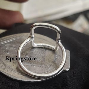 Peut inclure: Gros plan sur une bague argent&eacute;e avec un dessus rectangulaire et un anneau circulaire. La bague est pos&eacute;e sur une surface ovale argent&eacute;e avec le texte "Kpringstore". Le fond est noir.