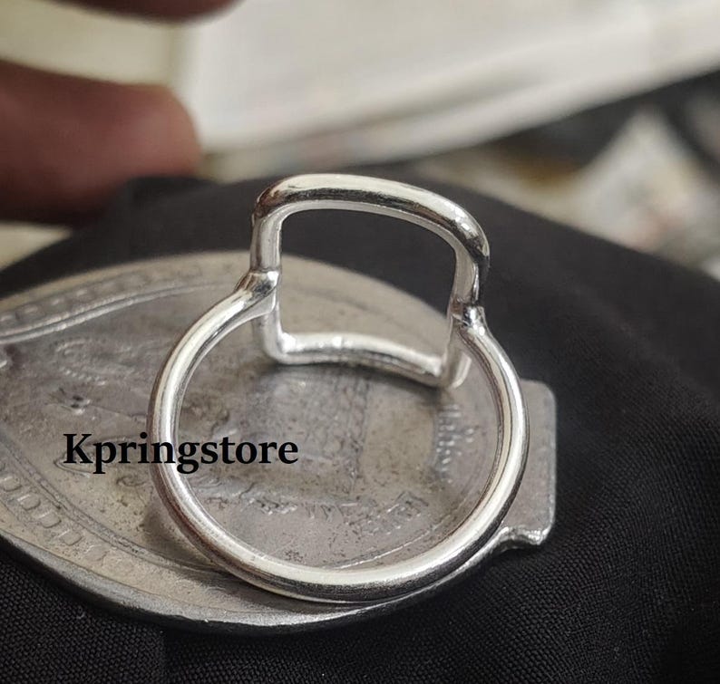 Peut inclure: Gros plan sur une bague argent&eacute;e avec un dessus rectangulaire et un anneau circulaire. La bague est pos&eacute;e sur une surface ovale argent&eacute;e avec le texte "Kpringstore". Le fond est noir.