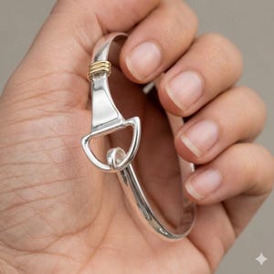 May include: A polished silver bangle bracelet featuring a stirrup-shaped focal point. The design includes a small, gold-toned metal accent consisting of three stacked rings positioned just above the stirrup element on the smooth silver band.