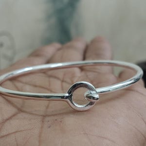 Op de afbeelding: Een zilverkleurige armband met een cirkelvormige sluiting. De armband heeft een glad, rond ontwerp. De sluiting is een kleine cirkel met een verbindingsstaaf. De armband wordt getoond op een hand. Diameter: ongeveer 6.5 cm.