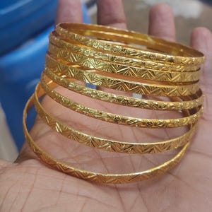 May include: A collection of gold bangles, each with detailed geometric and floral patterns. The bangles are stacked, showing their intricate designs and reflective surfaces. The jewellery is displayed on a hand.