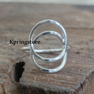 Może przedstawiać: Srebrny pierścionek z wzorem podwójnego koła. Pierścionek wykonany jest z cienkiego metalu i ma prosty, nowoczesny wygląd.