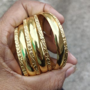 May include: Six gold bangles are displayed, each with a polished finish and a floral pattern along the edges. The bangles are held in a hand, reflecting light. The design is traditional, with a focus on the gold's shine and the intricate floral detailing. The bangles are a classic piece of jewellery.