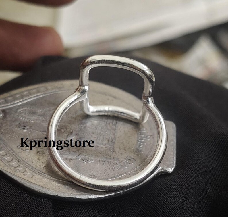 Peut inclure: Gros plan sur une bague argent&eacute;e avec un dessus rectangulaire et un anneau circulaire. La bague est pos&eacute;e sur une surface ovale argent&eacute;e avec le texte "Kpringstore". Le fond est noir.