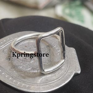 May include: A silver ring with a rectangular design. The ring is on a silver surface with the text "Kpringstore". The ring's design is a modern, minimalist style, with a square shape as the focal point.
