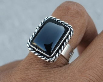 May include: A silver ring featuring a large, rectangular, polished black stone set in a frame with a twisted rope-style border. The band is smooth and metallic, showcasing a clean, reflective surface on the stone and intricate metal detailing.