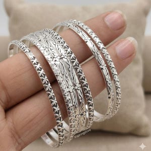 Peut inclure: Un ensemble de quatre bracelets joncs en métal argenté avec des motifs en relief complexes. Les bracelets présentent un design répétitif en forme d'étoile sur les bords et une bande centrale à motifs floraux. Le texte Kspringstore est visible.